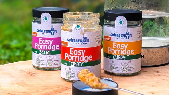 3 Easy Porridge Gläser auf Holztisch, eines geöffnet und zubereiteter Porridge auf einem Löffel davor