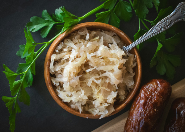 KI generiert: Das Bild zeigt eine Schale mit Sauerkraut und einem Löffel, daneben liegen zwei Bratwürste und frische Petersilie. Der Fokus liegt auf der appetitlichen Präsentation des Gerichts.