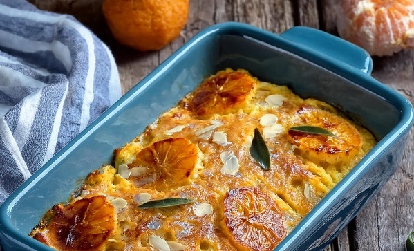 Das Bild zeigt ein gebackenes Gericht in einer blauen Auflaufform, garniert mit Orangenscheiben und Mandeln. Im Hintergrund liegen geschälte und ungeschälte Orangen sowie ein gestreiftes Geschirrtuch.
