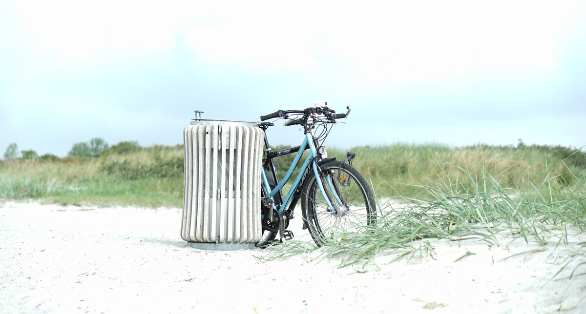 KI generiert: Ein Fahrrad lehnt an einem großen Mülleimer auf einem Sandweg, im Hintergrund sind Gräser.