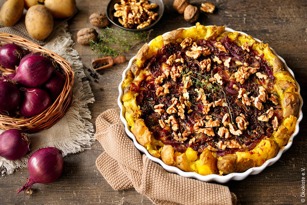 Das Bild zeigt eine herzhafte Tarte mit roten Zwiebeln und Walnüssen auf einem rustikalen Holztisch, umgeben von Zutaten wie roten Zwiebeln, Kartoffeln und Walnüssen. Die Tarte wird ansprechend präsentiert und deutet auf ein kulinarisches Thema hin.