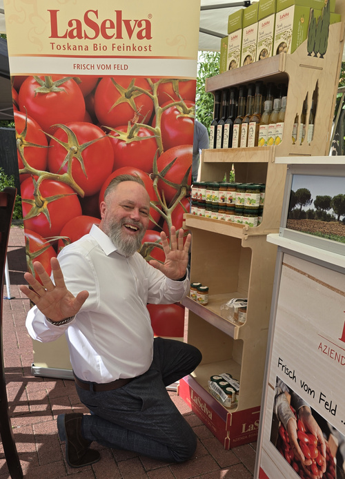 KI generiert: Ein Mann freut sich vor einem "LaSelva" Bio-Feinkoststand mit Tomatenmotiv. Text: "Frisch vom Feld".