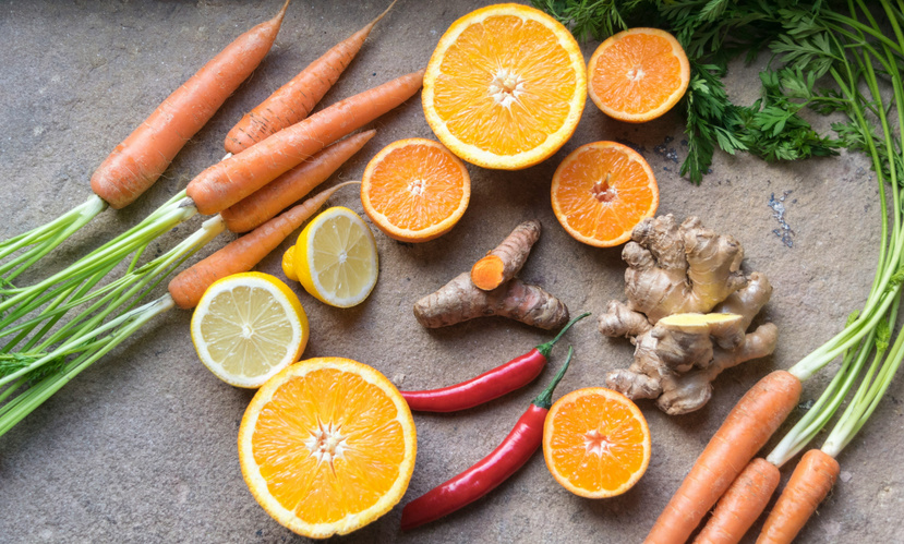 Das Bild zeigt eine Zusammenstellung frischer Zutaten: Karotten, Orangenscheiben, Zitronen, Chilischoten, Ingwer und Kurkuma auf einer Steinoberfläche. Der Fokus liegt auf der Vielfalt an Gemüse und Früchten.