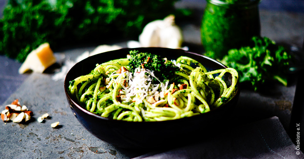 KI generiert: Eine Schüssel Spaghetti mit grünem Pesto, Nüssen und Parmesankäse. Text: „Ökokiste e. V.“