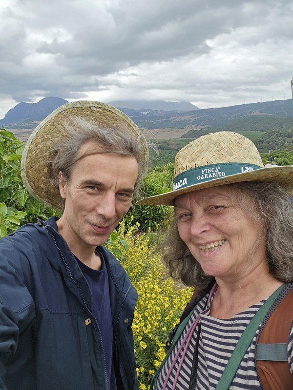 KI generiert: Zwei Personen mit Strohhüten lächeln in die Kamera, während sie in einer landschaftlichen Umgebung mit Bergen im Hintergrund stehen. Es ist bewölkt und im Vordergrund sind gelbe Blumen zu sehen.