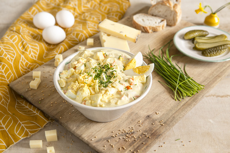Ein Eier-Käse-Salat in einer Schüssel auf einem Holzbrett mit Brot, Gurken und Frühlingszwiebeln.