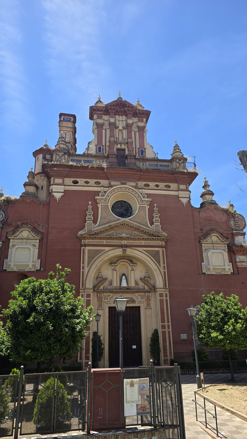 KI generiert: Rote Barockkirche mit verzierten Fassaden und einem runden Fenster, umgeben von Bäumen.