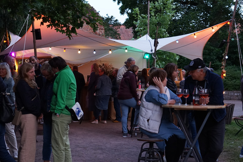 Das Bild zeigt eine gemütliche Outdoor-Veranstaltung mit mehreren Personen, die sich unter einem beleuchteten Pavillon unterhalten. Im Vordergrund sitzen zwei Personen an einem Stehtisch mit Getränken in Rotweingläsern.