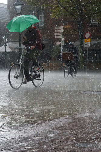 KI generiert: Zwei Radfahrer fahren im starken Regen; einer hält einen grünen Regenschirm. Im Hintergrund sind Geschäfte zu sehen.