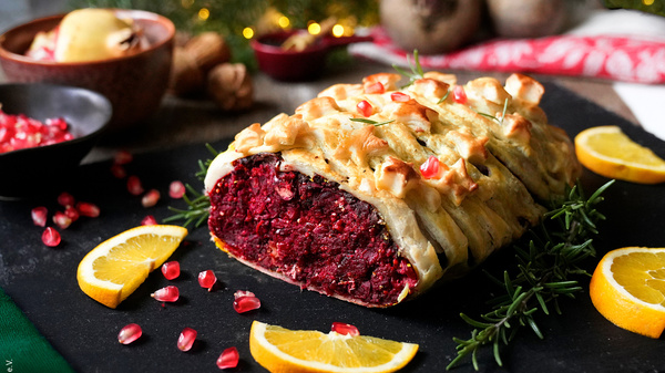 KI generiert: Das Bild zeigt einen mit Blätterteig umhüllten Rote-Bete-Braten, garniert mit Granatapfelkernen. Um das Gericht liegen frische Orangenscheiben und Kräuter.