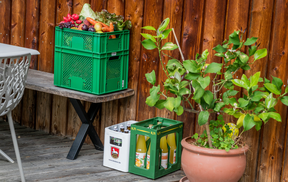 KI generiert: Grüne Kisten mit Obst, Gemüse und Saftflaschen stehen auf einem Tisch und Boden neben einer Pflanze.