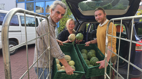 Drei Personen vor einem Lieferwagen mit Gemüsekisten voller Melonen.