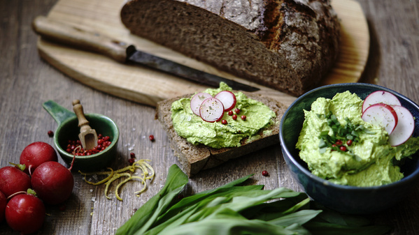 KI generiert: Brot mit grünem Aufstrich und Radieschen, daneben frische Radieschen und Bärlauch auf Holzbrett.
