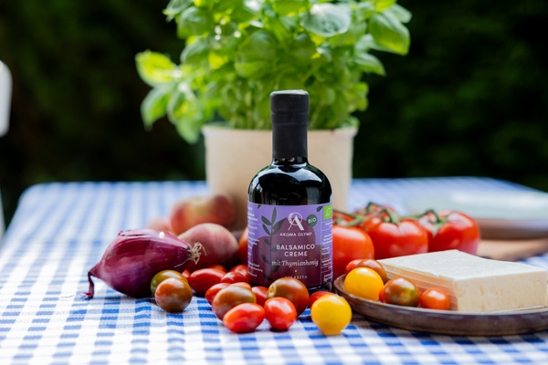 KI generiert: Flasche Balsamico-Creme, Gemüse und Feta auf blau-weiß kariertem Tisch, Basilikum im Hintergrund. Text: "BALSAMICO CREME".