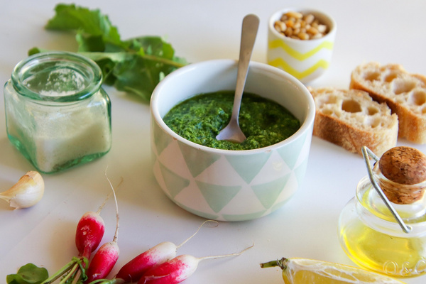 KI generiert: Eine Schale mit grünem Pesto, umgeben von Brot, Öl, Radieschen, Knoblauch und Käse.