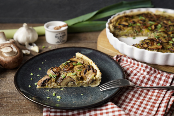 KI generiert: Ein Stück Pilzquiche auf einem Teller mit Gabel, Knoblauch und Lauch im Hintergrund.
