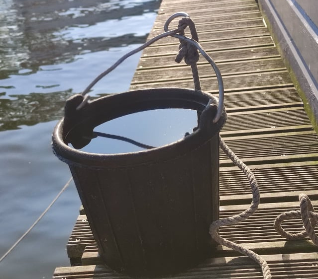 KI generiert: Ein schwarzer Eimer voller Wasser auf einem Holzsteg am Wasser.