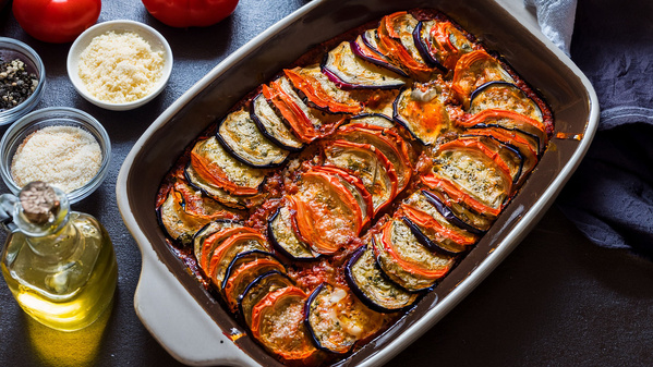 Ein Auflauf aus Tomaten, Zucchini und Auberginen in einer Keramikform, daneben Zutaten wie Öl und Parmesan.