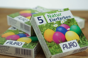 KI generiert: Verpackung von AURO Natur-Eierfarben für fünf bunte Eier auf einer Wiese.