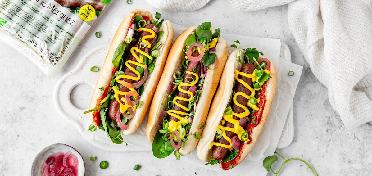 KI generiert: Drei vegane Hotdogs mit Senf, grünen Blättern und Gemüse. Verpackungstext: "Vegane Merguez".