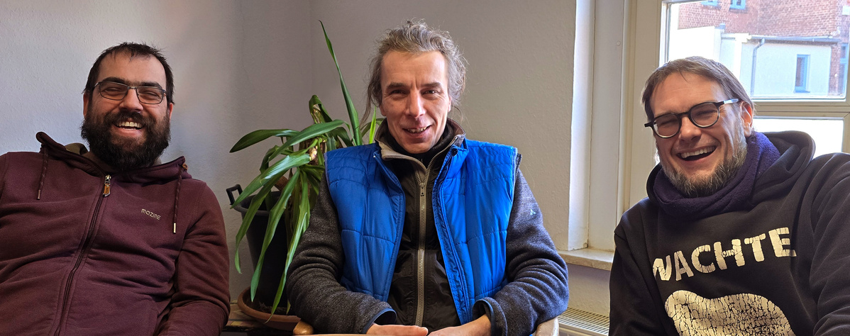 KI generiert: Drei lächelnde Männer sitzen drinnen; einer trägt einen "WACHTET"-Pullover.