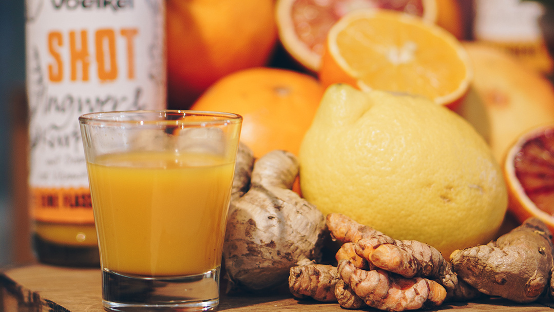 Voelkel Ingwershot im Glas umgeben von Ingwer, Kurkuma, Zitrusfrüchten