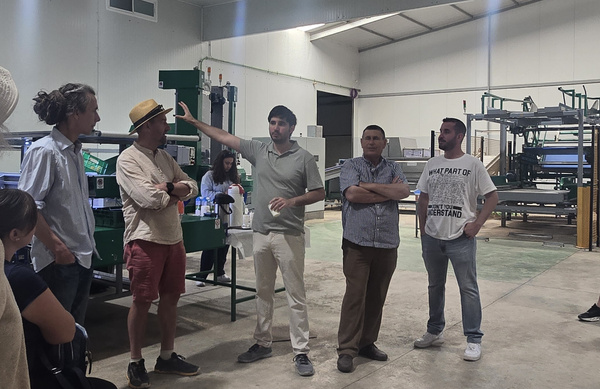 KI generiert: Eine Gruppe von Menschen steht in einer Fabrikhalle. Ein Mann erklärt etwas. Text auf Shirt: "WHAT PART OF ...".