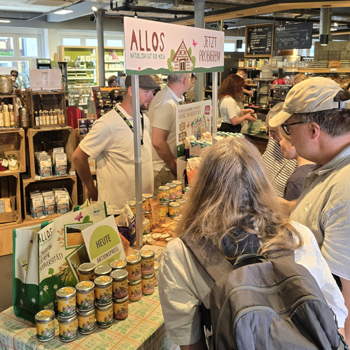 KI generiert: Menschen an einem Probierstand der Marke Allos in einem Lebensmittelladen. Text: "Jetzt probieren!"