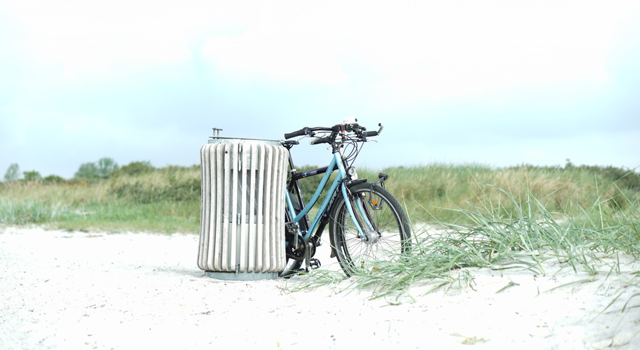 Ein Fahrrad lehnt an einem Mülleimer am Strand mit Dünengras im Hintergrund.