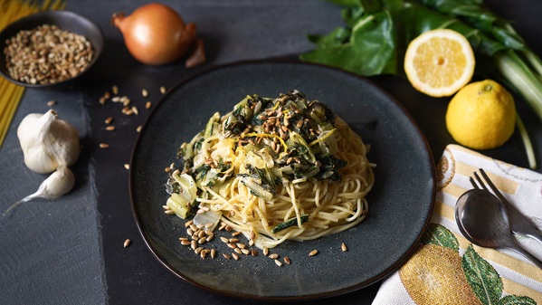 Ein Teller Spaghetti mit Mangold und gerösteten Kernen, umgeben von Zwiebel, Knoblauch und Zitrone.