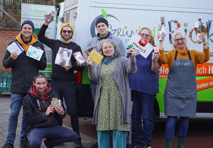 Eine Gruppe von Menschen steht fröhlich vor einem Lieferwagen und hält Geschenke und Umschläge in den Händen. Sie scheinen für eine Feier oder Aktion zusammenzukommen.