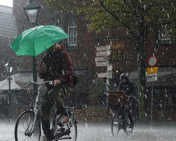 KI generiert: Zwei Personen fahren bei starkem Regen Fahrrad, einer hält einen grünen Regenschirm.