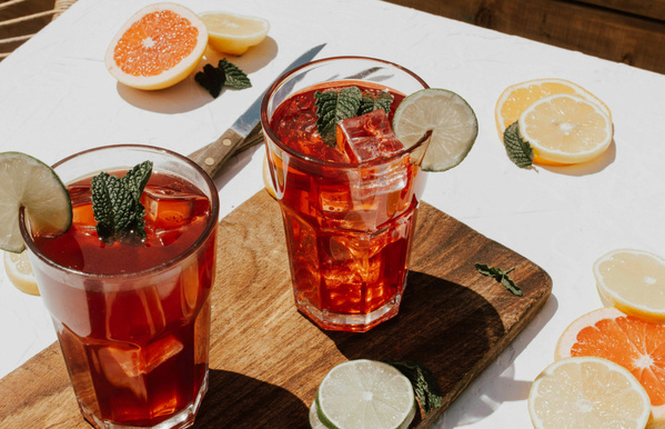 KI generiert: Zwei rote Getränke mit Eis, Limette und Minze auf einem Holzbrett, umgeben von Zitrusfrüchten.