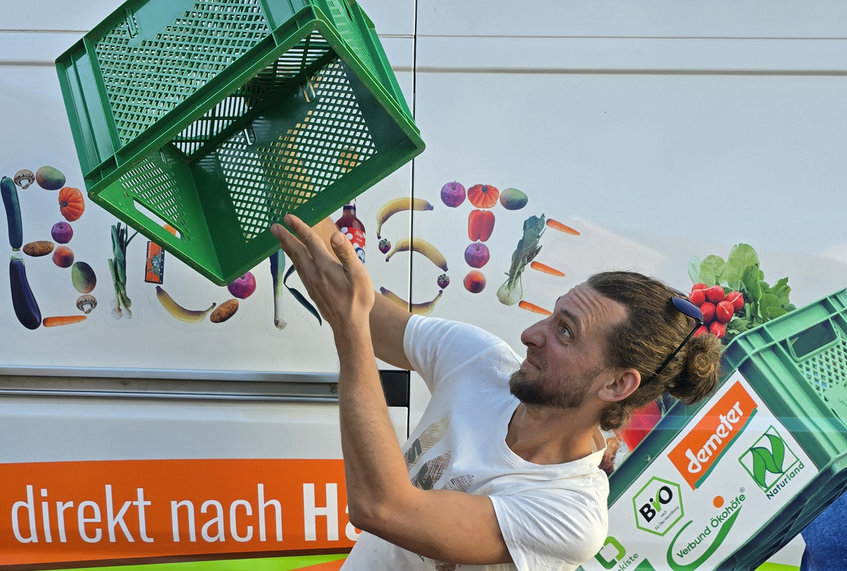 KI generiert: Ein Mann balanciert eine grüne Kiste vor einem Lieferwagen mit Gemüsewerbung. Text: "direkt nach H".