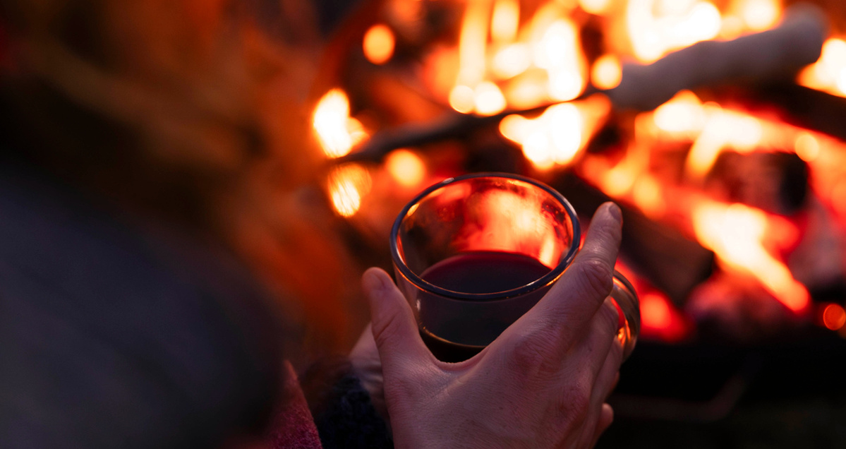 Das Bild zeigt eine Person, die eine Tasse wärmt, während im Hintergrund ein Lagerfeuer lodert. Der Fokus liegt auf der gemütlichen Atmosphäre und der Wärme des Feuers.