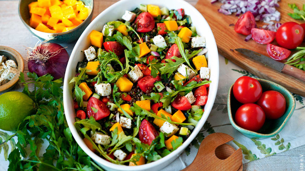 Frischer Salat mit Tomaten, Rucola, Mango und Feta in einer Schüssel, umgeben von vorbereiteten Zutaten.