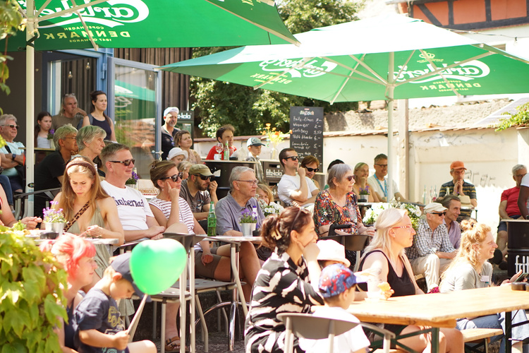 KI generiert: Eine Gruppe von Menschen sitzt draußen unter grünen Sonnenschirmen und schaut einem Ereignis zu.