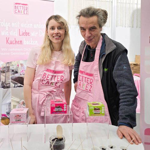 KI generiert: Zwei Personen in rosa Schürzen präsentieren Kuchen; Text: „… wie viel Liebe wir Deine Kuchen backen.“