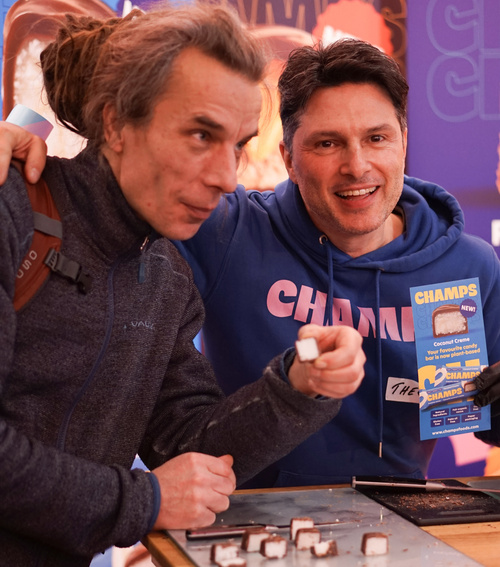 KI generiert: Zwei Männer probieren Süßigkeiten, einer hält eine Broschüre mit „CHAMPS“ und „Coconut Crème“.