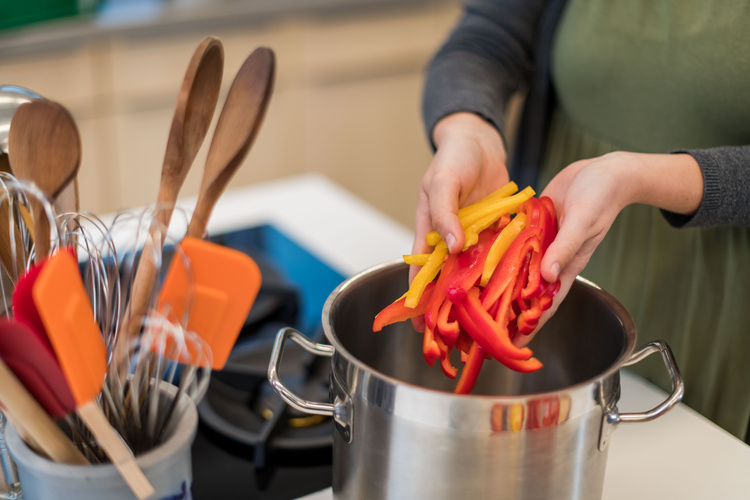 Köchin gibt Paprikastreifen gelb und rot in hohen Edelstahltopf, links daneben steht ein Steinguttopf mit Küchenutensilien