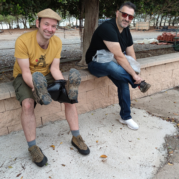 KI generiert: Das Bild zeigt zwei lachende Männer, die auf einer niedrigen Mauer sitzen und ihre Schuhe mit improvisierten Abdeckungen aus Plastiktüten versehen haben. Im Hintergrund ist eine lockere Baumkulisse zu sehen.