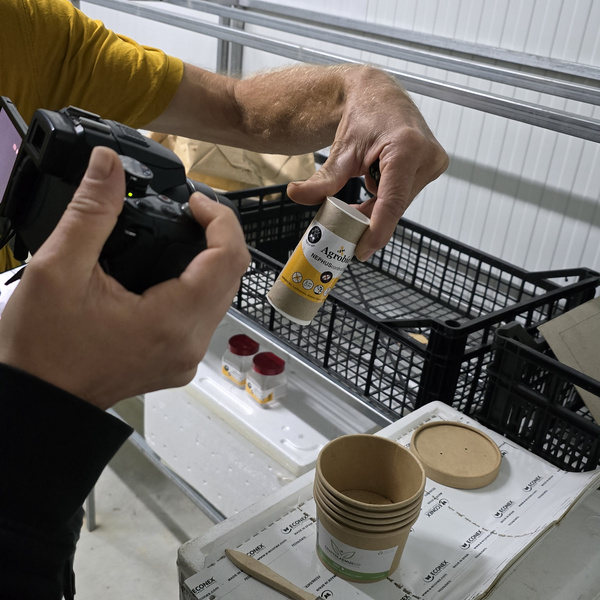 KI generiert: Das Bild zeigt eine Person, die eine Flasche Novizym in den Händen hält, während eine andere Person sie fotografiert. Im Hintergrund sind leere Becher und eine schwarze Kiste zu sehen.