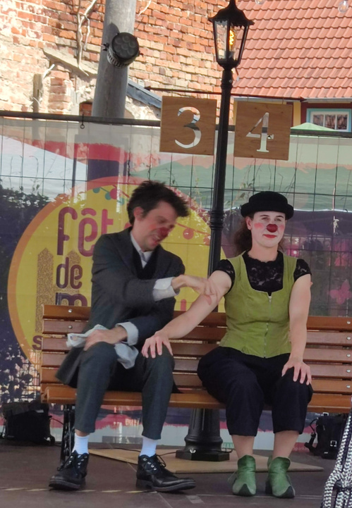 KI generiert: Zwei Clowns performen auf einer Bühne mit einer Laterne und einer Bank. Hintergrundtext: "fête de".