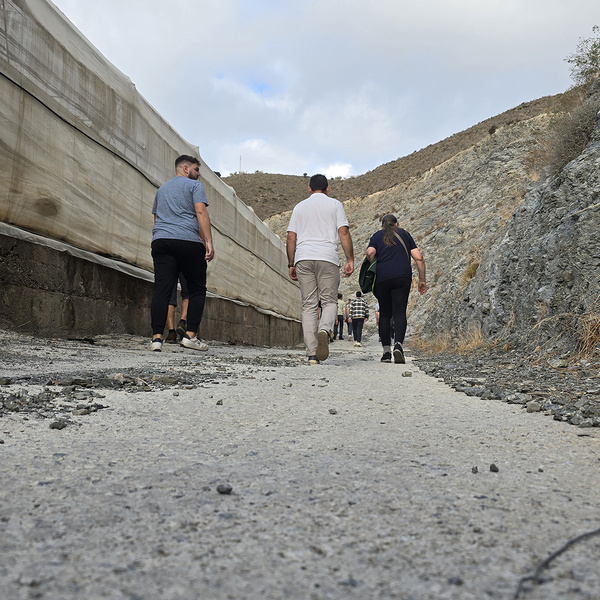 KI generiert: Eine Gruppe von Menschen geht auf einem steinigen Weg, umgeben von felsigen Hängen und einem Bauwerk mit einer Plane auf der linken Seite. Das Bild vermittelt das Gefühl einer Wanderung oder Erkundung in einer kargen Landschaft.