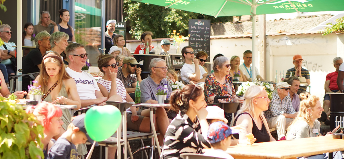 Menschen sitzen im Freien, einige mit Getränken, und genießen ein Event bei schönem Wetter.