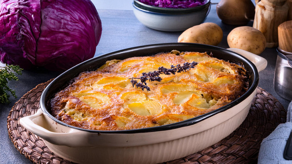 KI generiert: Das Bild zeigt einen frisch gebackenen Kartoffelauflauf in einer weißen Auflaufform, garniert mit einem Zweig Kräuter. Im Hintergrund sind rote Kohlköpfe und Kartoffeln zu sehen.