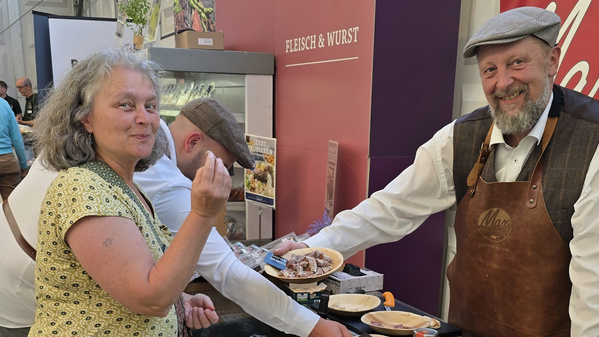 KI generiert: Ein Mann bietet einer Frau Wurstproben an einem Messestand an. Text: "FLEISCH & WURST".