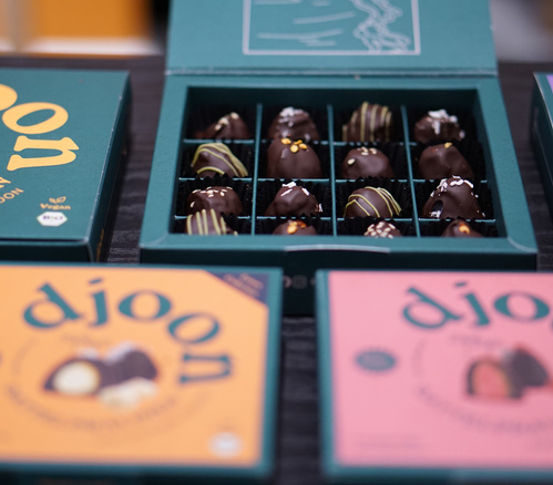 KI generiert: Eine Schachtel mit Pralinen, umgeben von bunten Verpackungen. Text: "djoon".