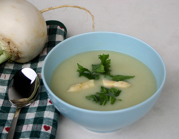 KI generiert: Eine Schüssel mit cremiger Suppe, garniert mit Spargel und Kräutern, daneben ein großer Rettich und Löffel auf Tischdecke.