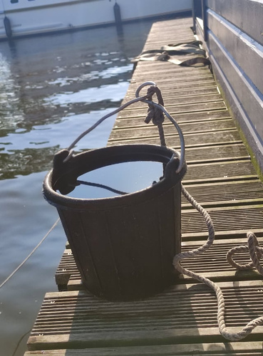 KI generiert: Ein schwarzer Plastikeimer mit Seil steht auf einem Steg am Wasser.